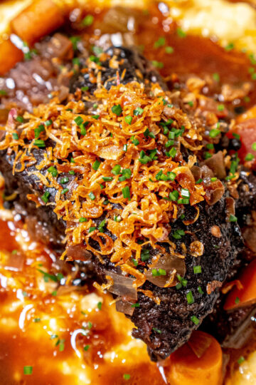 A close-up of braised beef short rib topped with crispy fried onions and chopped chives, served over creamy mashed potatoes with gravy and pieces of carrots.