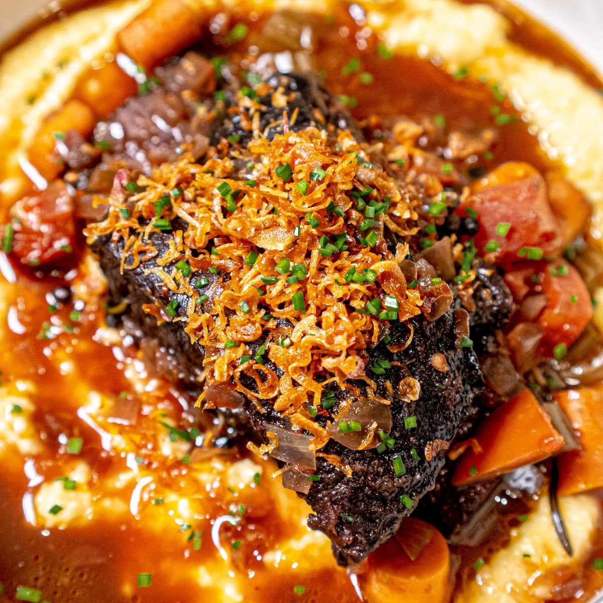 A close-up of braised beef short rib topped with crispy fried onions and chopped chives, served over creamy mashed potatoes with gravy and pieces of carrots.