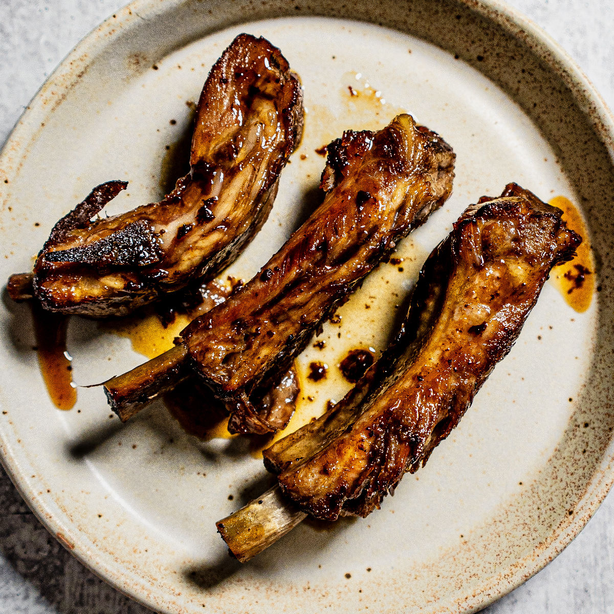 Three cooked pork ribs with a caramelized glaze are arranged on a rustic beige plate, with some sauce drizzled around them. The ribs have a slightly charred appearance.