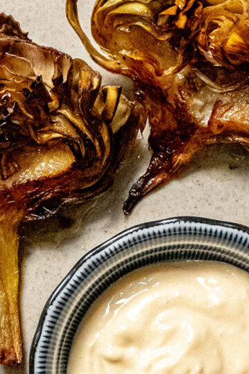 Two crispy, roasted artichoke halves on a plate next to a small bowl of creamy dipping sauce. The artichokes have golden-brown edges and are arranged on a neutral-toned surface.