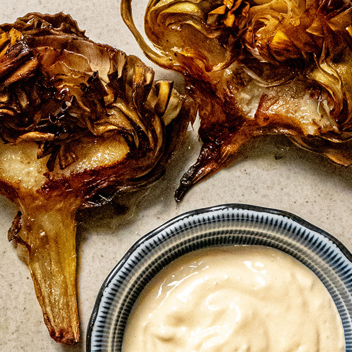 Two crispy, roasted artichoke halves on a plate next to a small bowl of creamy dipping sauce. The artichokes have golden-brown edges and are arranged on a neutral-toned surface.
