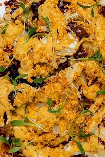 Close-up of roasted cauliflower florets topped with a creamy orange sauce, crunchy golden crumbs, and garnished with fresh green microgreens on a white plate.