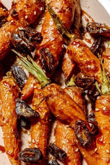 A platter of roasted carrots topped with a glossy sauce and garnished with chopped dark olives and herbs, served on a light-colored dish.