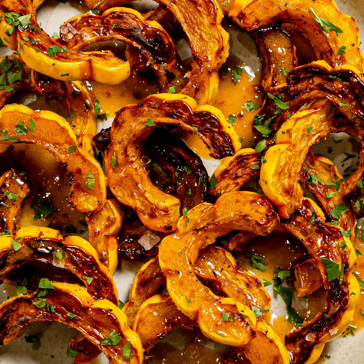 Slices of roasted delicata squash with caramelized edges, drizzled with a glossy sauce and sprinkled with fresh chopped herbs on a light-colored surface.