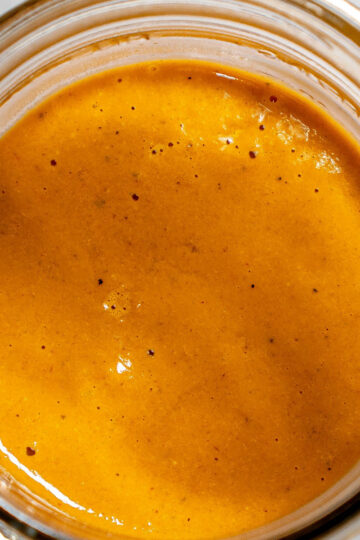 A close-up, top-down view of a glass jar filled with thick, smooth, orange-brown sauce with small black specks.