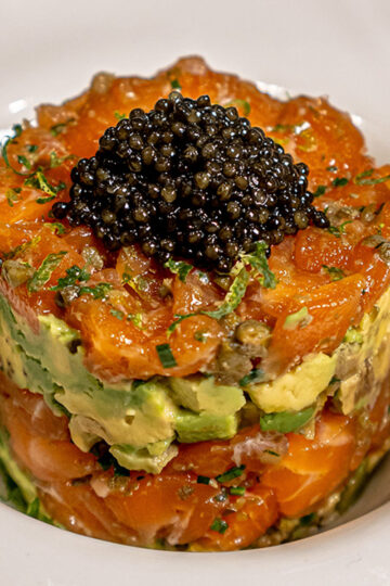 A gourmet dish featuring a stacked salmon and avocado tartare, garnished with a generous serving of black caviar on top, presented in a white bowl.
