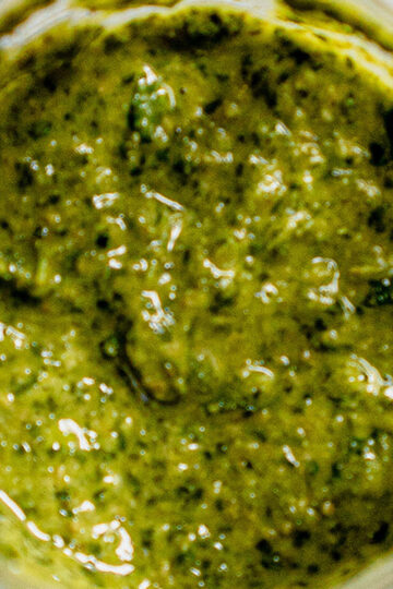 Close-up view of a jar filled with green pesto sauce. The sauce has a textured, slightly chunky appearance with visible flecks of herbs and oil.