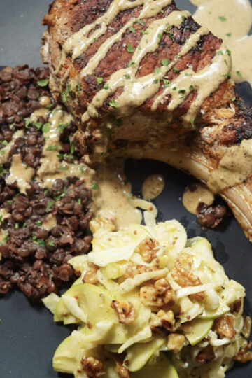 A plate with a grilled pork chop topped with creamy sauce, served with cooked lentils and a side of salad made with cabbage, green apples, and walnuts.