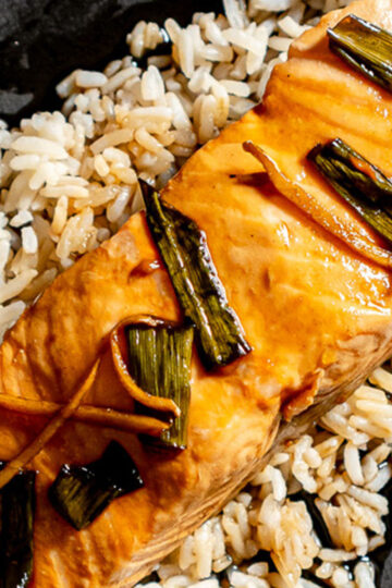 A close-up of a cooked salmon fillet topped with green onions and ginger, served on a bed of brown rice.
