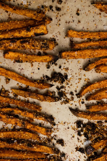 Close-up of baked sweet potato fries scattered on a baking sheet, coated with spices and seasoning, with bits of seasoning spread around.