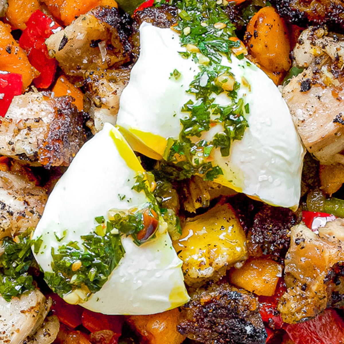 A close-up of a colorful hash with roasted vegetables and crispy meat, topped with two poached eggs drizzled with green herb sauce. The eggs are slightly broken, showing the soft yolk.