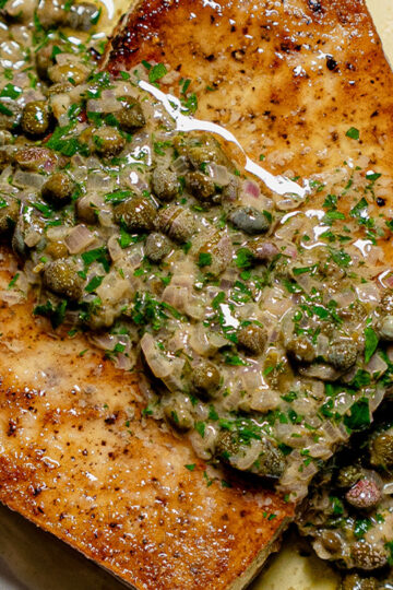 A close-up of two golden-brown fish fillets topped with a creamy sauce containing capers, herbs, and finely chopped onions.