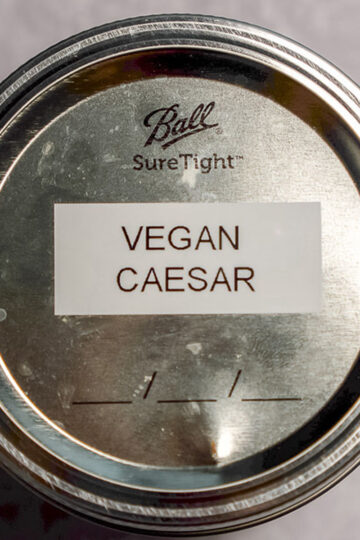 A metal jar lid labeled Vegan Caesar with Ball SureTight stamped on the top, viewed from above against a light background.