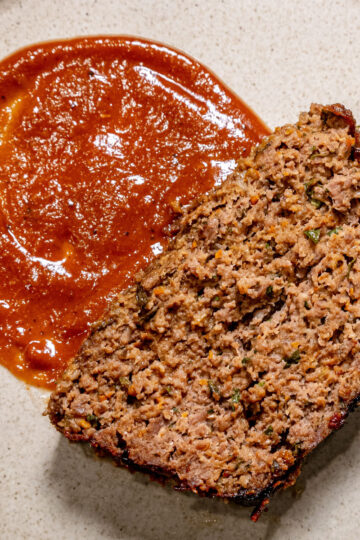 A slice of meatloaf sits on a beige plate next to a serving of red tomato sauce. The meatloaf appears moist and seasoned, with visible bits of herbs or vegetables.