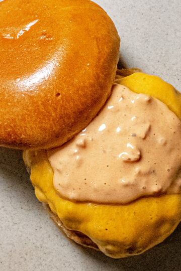 A close-up of an open cheeseburger with a shiny top bun, melted cheddar cheese, and a generous layer of creamy sauce on the patty, served on a light-colored plate.