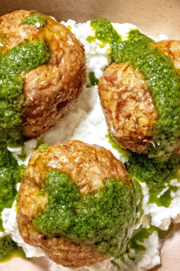 Three browned meatballs sit on a bed of white ricotta cheese, topped with a drizzle of vibrant green pesto sauce.