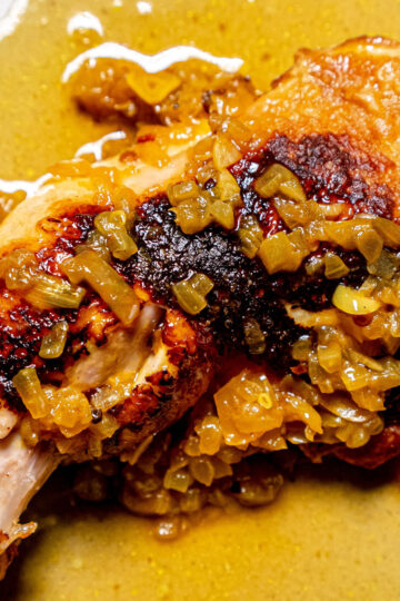 A close-up of a cooked chicken leg topped with a chunky, golden-brown sauce made of onions and spices, served with its juices on a white plate.