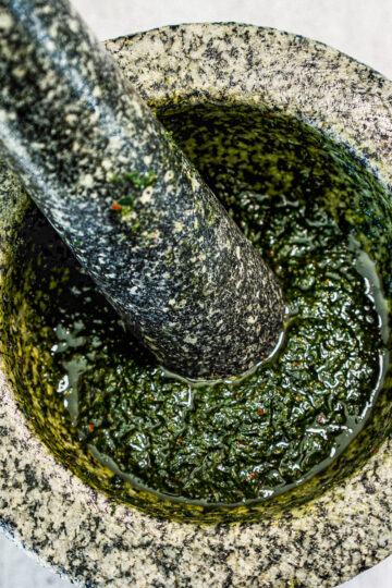 A close-up of a stone mortar and pestle with green pesto being ground inside, showing a coarse texture and oily surface.