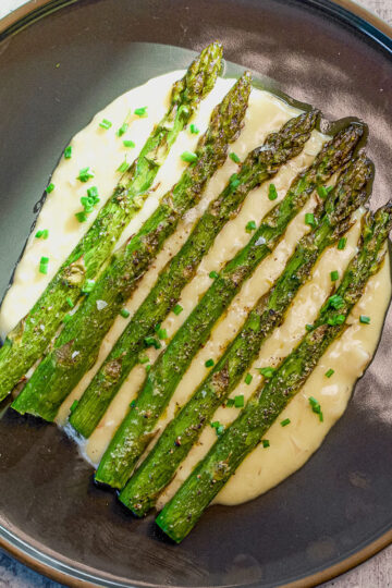 Six roasted asparagus spears are arranged on a bed of creamy mashed potatoes, garnished with chopped chives, served on a dark plate.
