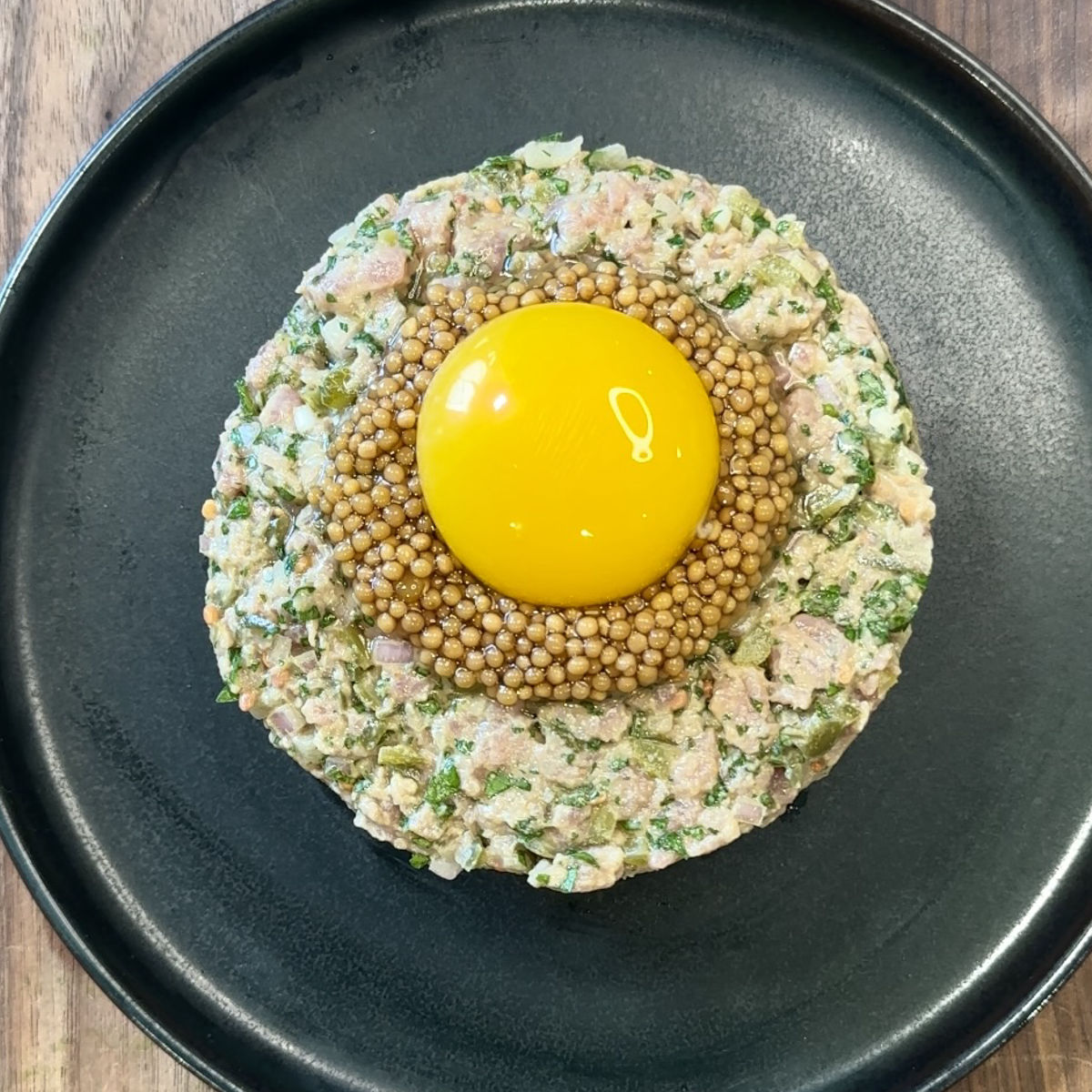 A round serving of steak tartare topped with a layer of mustard seeds and a raw egg yolk, arranged neatly on a black plate.