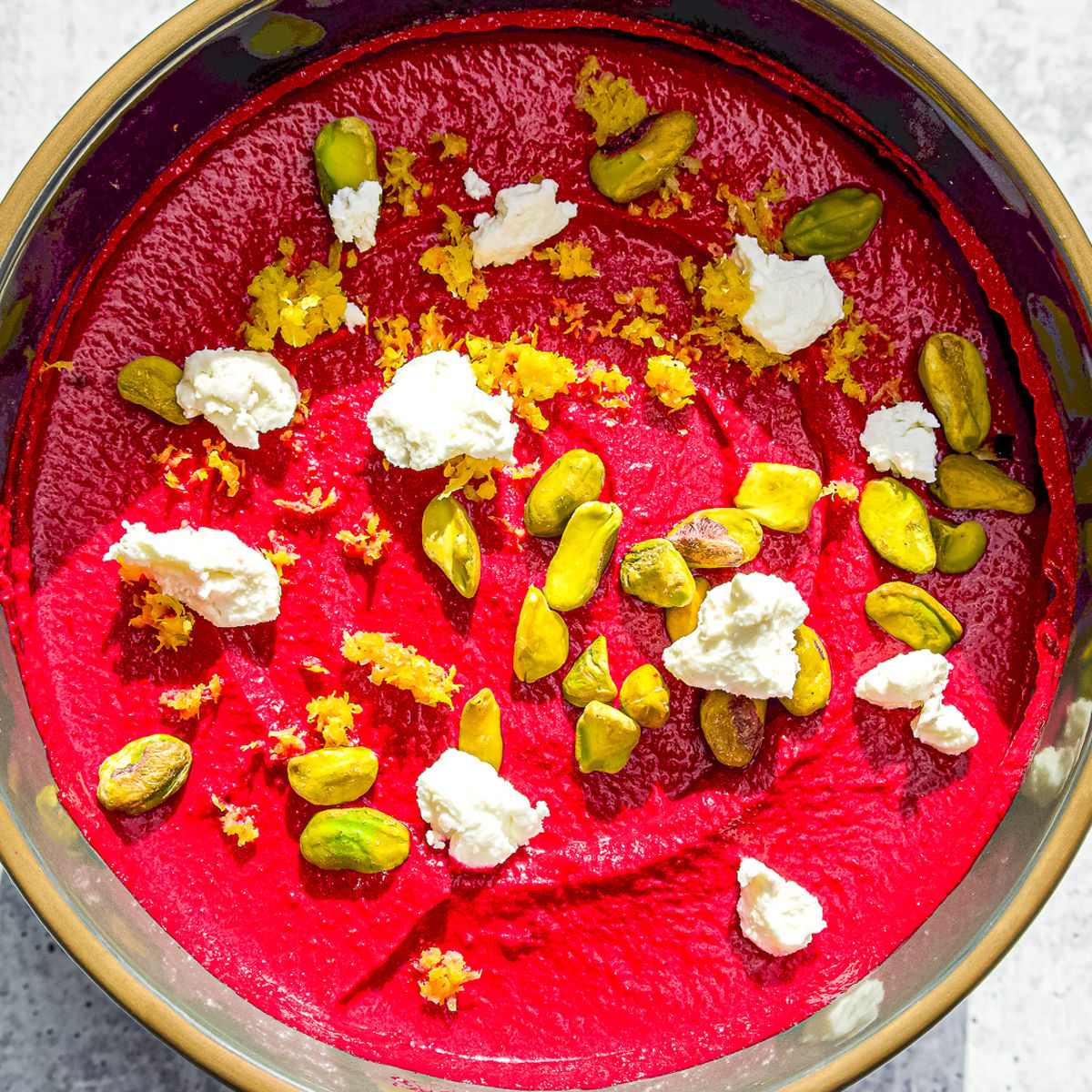 A bowl of vibrant beet hummus topped with whole pistachios, dollops of white goat cheese, and sprinkled with lemon zest. The bowl sits on a light, textured surface.