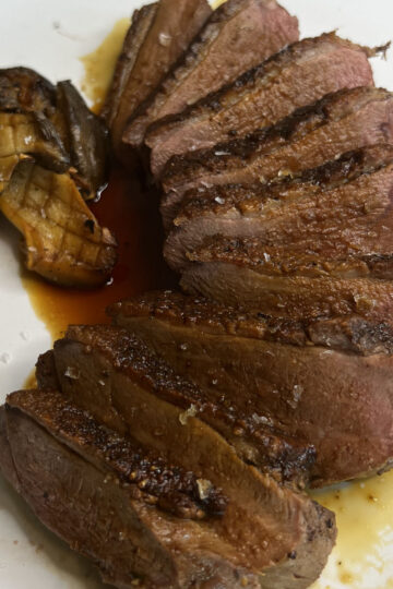 Sliced seared steak arranged on a white plate with grilled mushrooms on the side, drizzled with a light brown sauce.