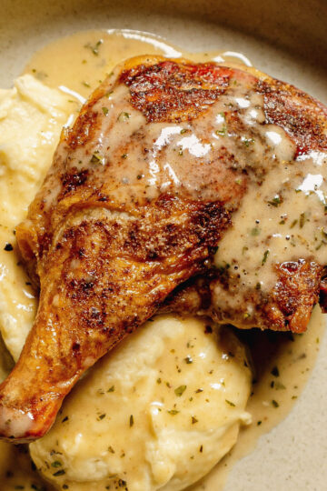 A roasted chicken leg with crispy skin sits atop creamy mashed potatoes, all drizzled with a rich, herb-speckled gravy on a beige plate.