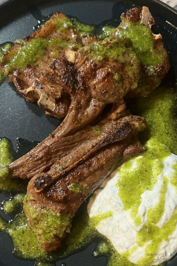 Three grilled lamb chops on a black plate, drizzled with green chimichurri sauce, served alongside a generous portion of creamy white dip or sauce.