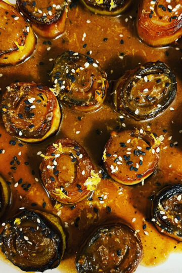 Close-up of roasted leeks cut into rounds, topped with a glossy reddish-brown sauce and sprinkled with black and white sesame seeds on a white plate.