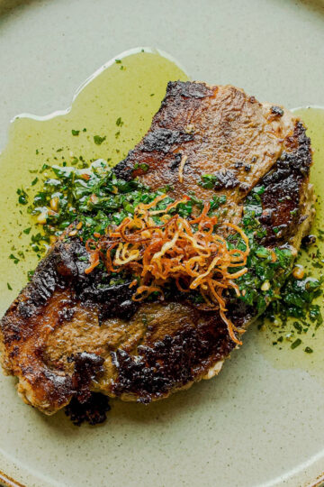 A seared steak sits on a light green plate, topped with crispy fried onions and fresh chopped herbs, surrounded by a drizzle of olive oil.