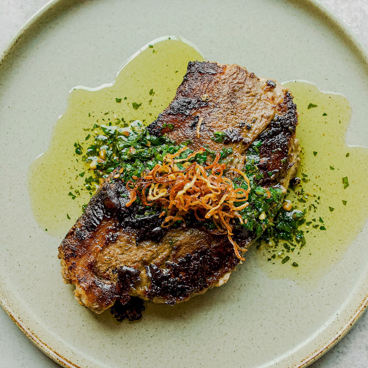 A seared steak sits on a light green plate, topped with crispy fried onions and fresh chopped herbs, surrounded by a drizzle of olive oil.