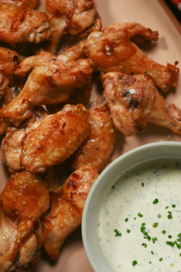 A platter of baked chicken wings is served next to a bowl of creamy ranch dip garnished with chopped chives. A can of Dr Pepper soda is partially visible in the corner.