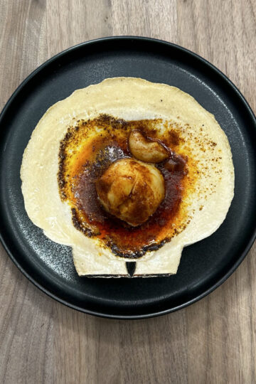 A black plate holds a tortilla topped with a round ball of food, possibly a potato, roasted garlic cloves, and a pool of reddish-brown sauce, all set on a light wood surface.