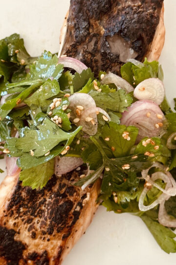 A piece of grilled salmon topped with a fresh herb salad made of cilantro, sliced shallots, and sesame seeds, served on a white plate.