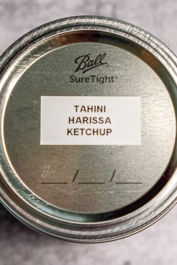 A metal mason jar lid labeled TAHINI HARISSA KETCHUP sits on a gray textured surface. The lid is seen from above, showing the Ball SureTight brand and a white label with the sauces name.