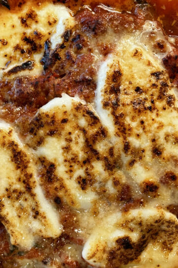 Close-up of baked lasagna showing browned, melted cheese on top of a rich tomato meat sauce, with a bubbling, golden crust.
