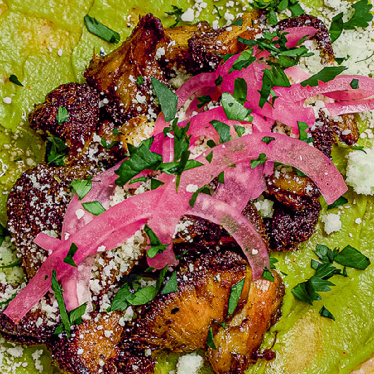 Close-up of seasoned grilled chicken pieces topped with pickled red onions, crumbled cheese, and chopped fresh herbs, served on a layer of green sauce or guacamole.