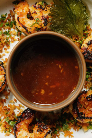 Grilled shrimp arranged in a circle on a plate with crispy herbs and crumbs, surrounding a bowl of dark dipping sauce in the center. A single leafy green garnish decorates the edge of the plate.