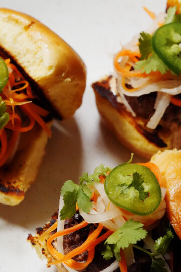 Three small sliders with grilled meat, pickled carrots and daikon, fresh cilantro, and sliced jalapeños on toasted buns, arranged on a white plate.
