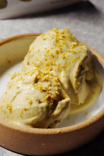 Two scoops of creamy ice cream topped with yellow zest are served in a round, beige ceramic bowl on a gray surface.