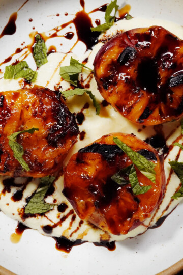 Three caramelized peach halves are served on a bed of creamy mascarpone, drizzled with balsamic glaze and garnished with fresh mint leaves on a white plate with a brown rim.
