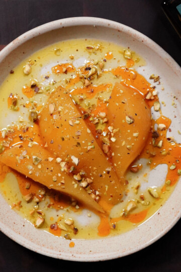 Three slices of poached pear are arranged on a beige plate, topped with chopped pistachios and drizzled with golden syrup and orange sauce. A green-labeled bottle appears partially in the upper right corner.