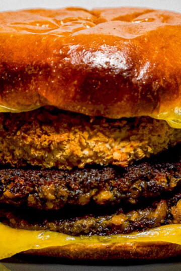 A close-up of a large burger with a shiny brioche bun, two crispy, charred veggie patties, melted yellow cheese, and a breaded, fried patty in the middle, all stacked on a dark plate.