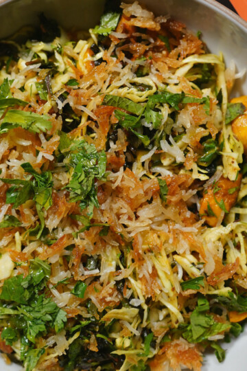 A close-up of a bowl of fresh salad with shredded cabbage, fried crispy toppings, chopped green herbs, and yellow vegetables, garnished with cilantro.
