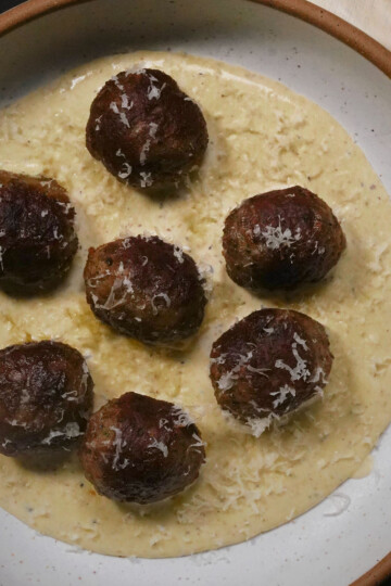 Seven browned meatballs sit on a creamy white sauce, garnished with grated cheese, served on a white plate with a brown rim.