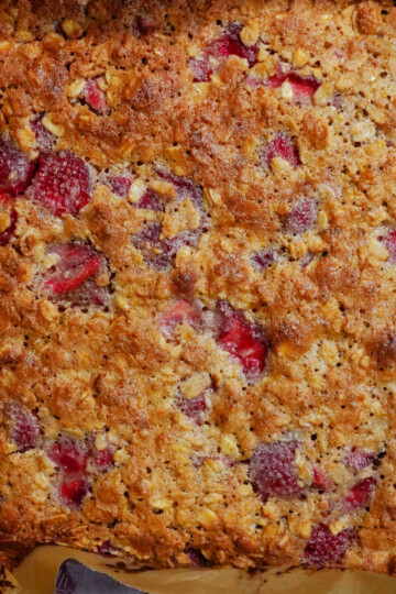A close-up of a freshly baked strawberry oat bar in a baking dish, showing a golden-brown crust with visible oats and chunks of strawberries throughout.