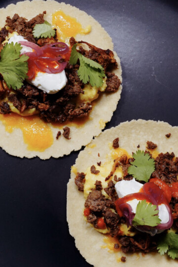 Two corn tortillas topped with seasoned ground meat, cheese sauce, sour cream, pickled red onions, hot sauce, and fresh cilantro are arranged on a dark plate.