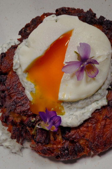 A crispy potato rösti topped with a poached egg, split open to reveal a runny yolk, garnished with edible purple flowers and served with a dollop of creamy herb sauce on a white plate.