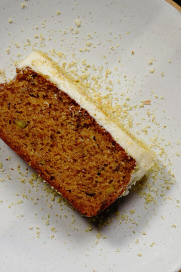 A slice of moist carrot cake with cream cheese frosting sits on a white plate, lightly sprinkled with grated zest or crumbs.