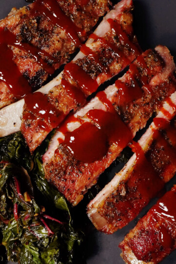 Sliced barbecue ribs drizzled with sauce on a dark plate, served with a side of sautéed greens.